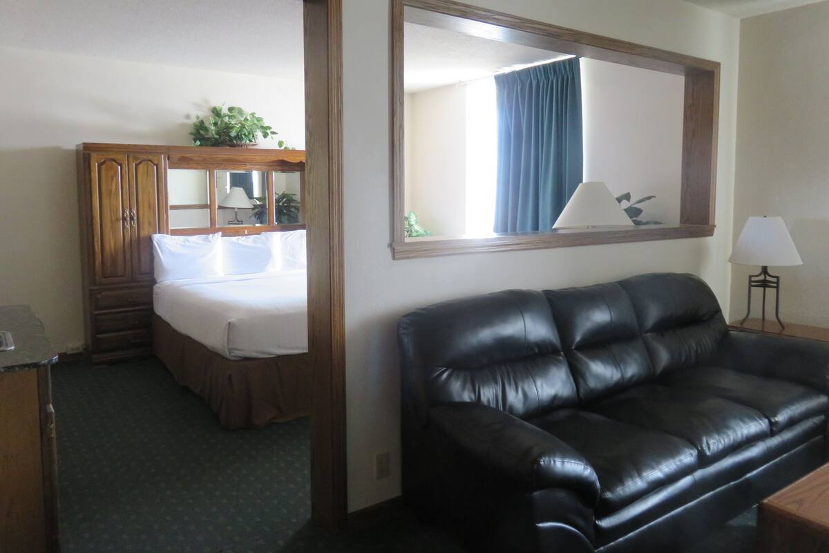 A hotel room with a bed, a black leather sofa, a wooden wardrobe, a mirror, and a lamp.