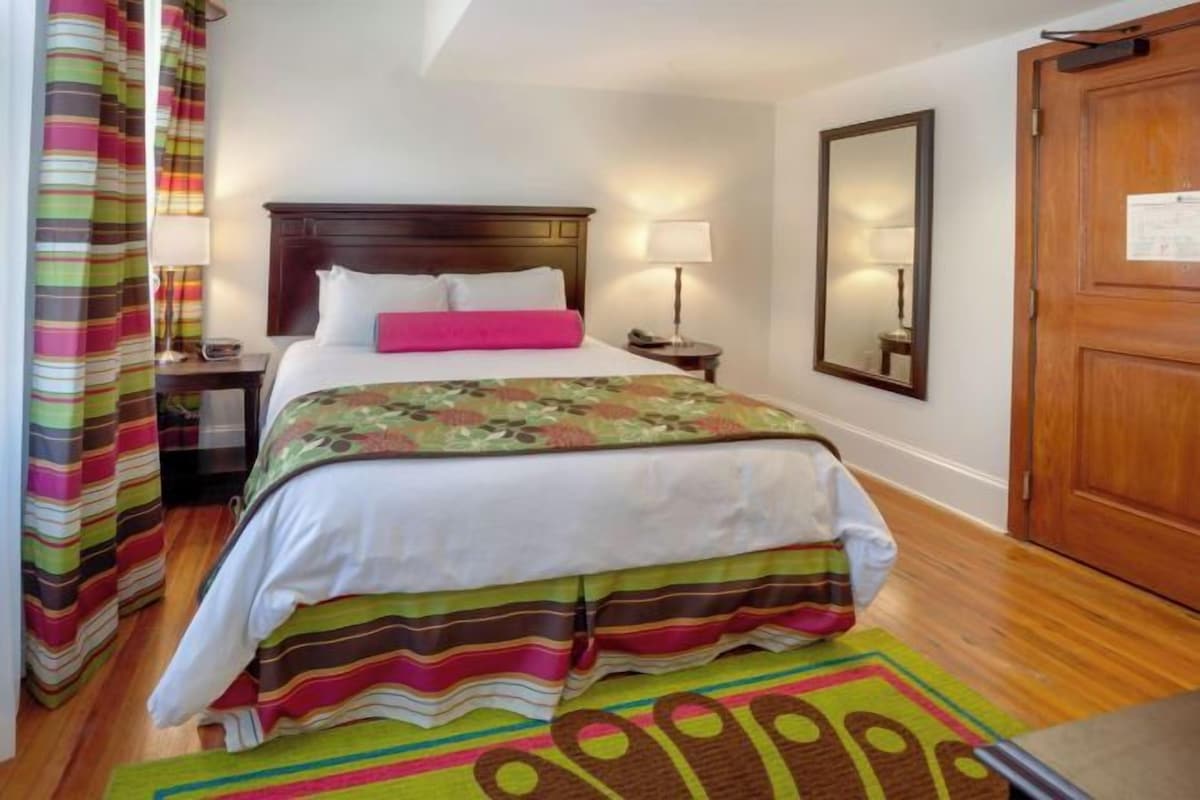 A hotel room with a bed, a nightstand, a lamp, a mirror, and a wooden door.