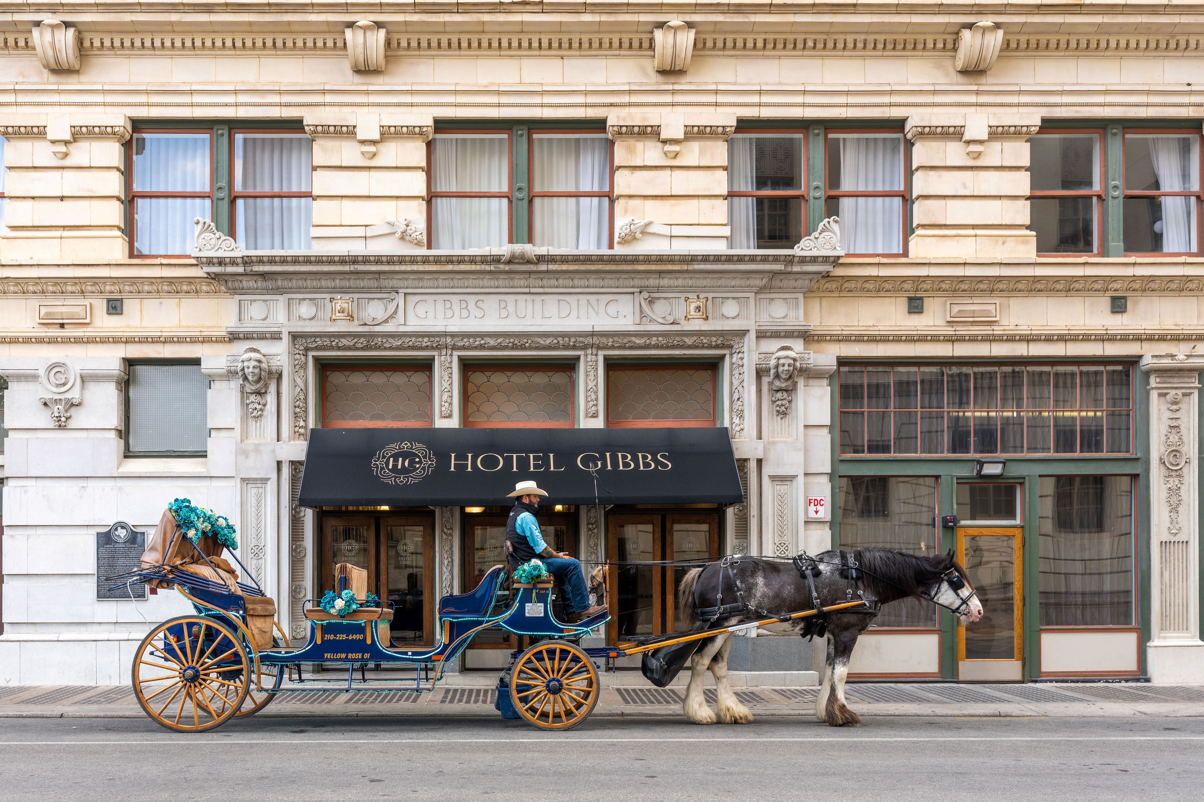 Une calèche tirée par des chevaux se trouve devant un bâtiment sur lequel on peut lire l’inscription « HÔTEL GIBBS ».