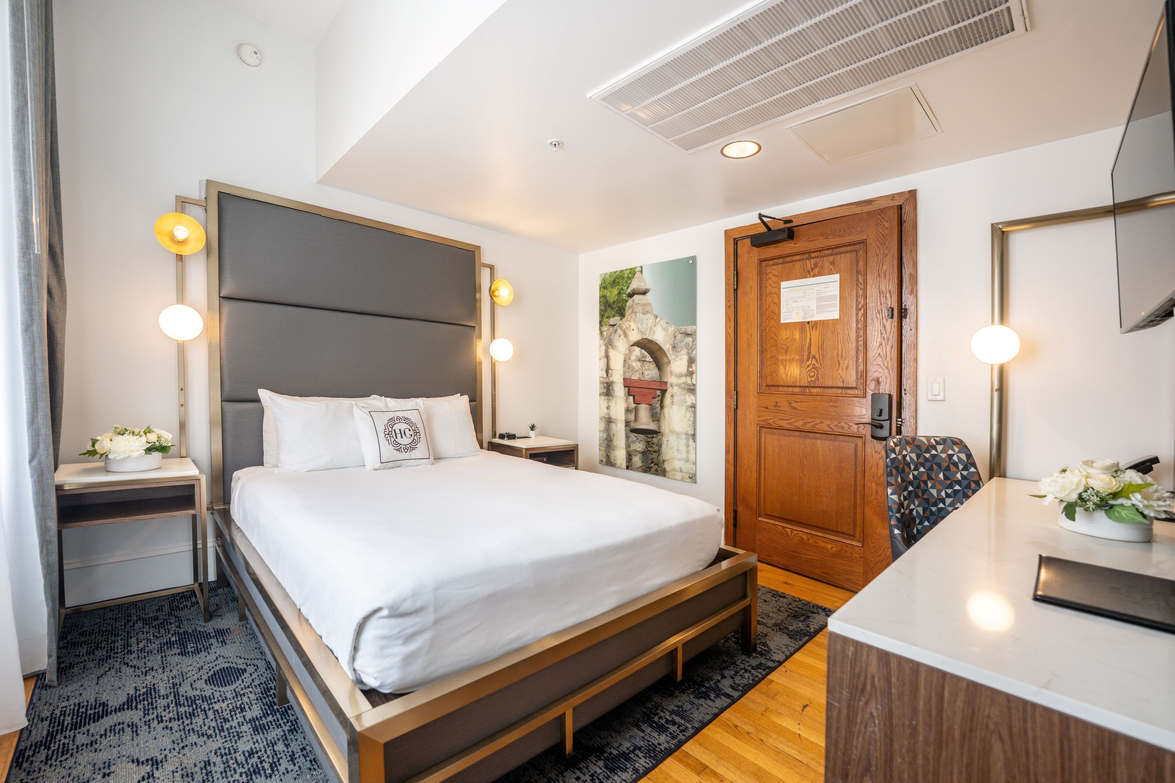A modern hotel room with a large bed, bedside tables, a desk, and a door with a wooden panel.