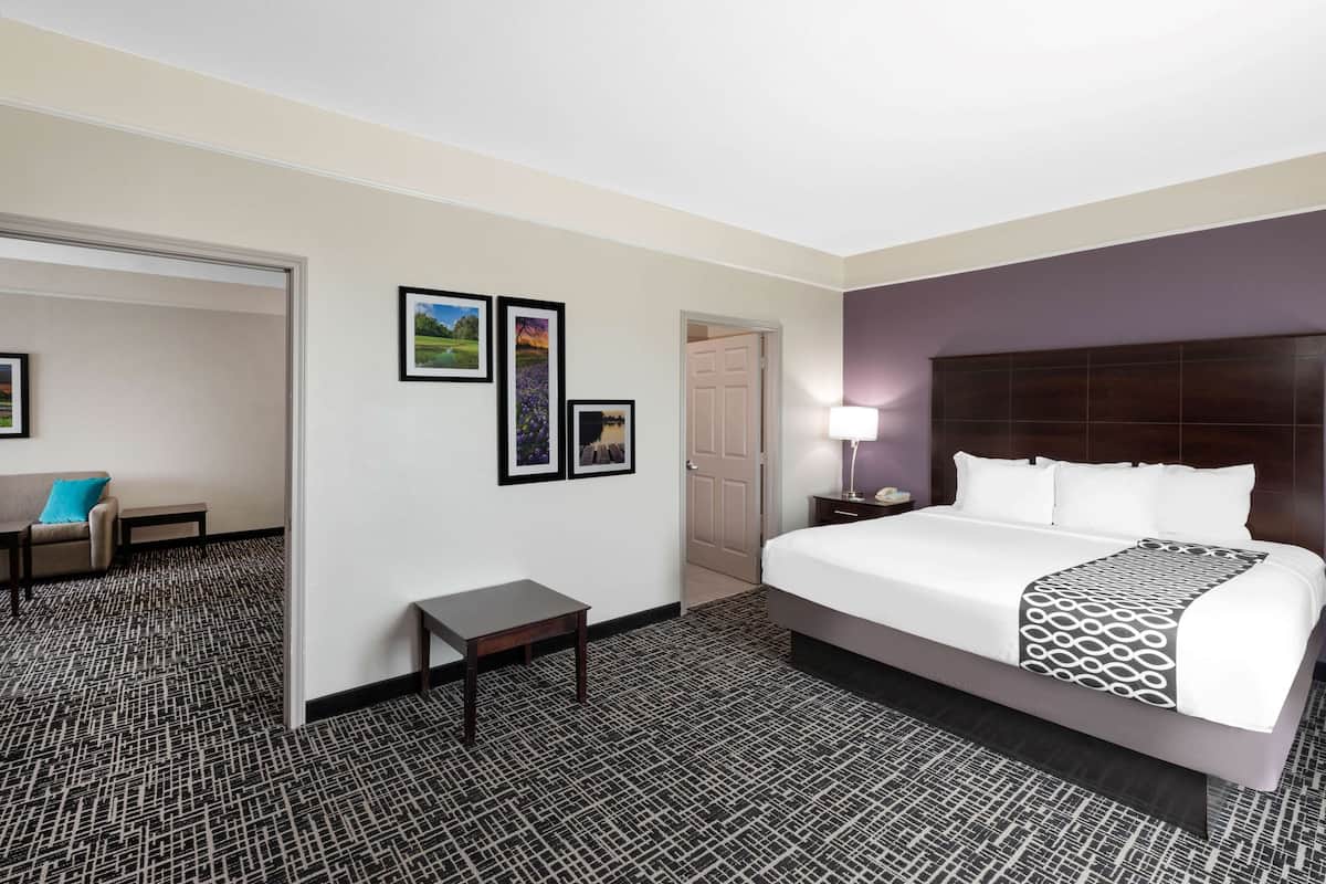 Habitación de hotel con una cama grande, una mesita de noche, un escritorio y una silla. Se observan cuadros en la pared y una puerta que da a otra habitación.