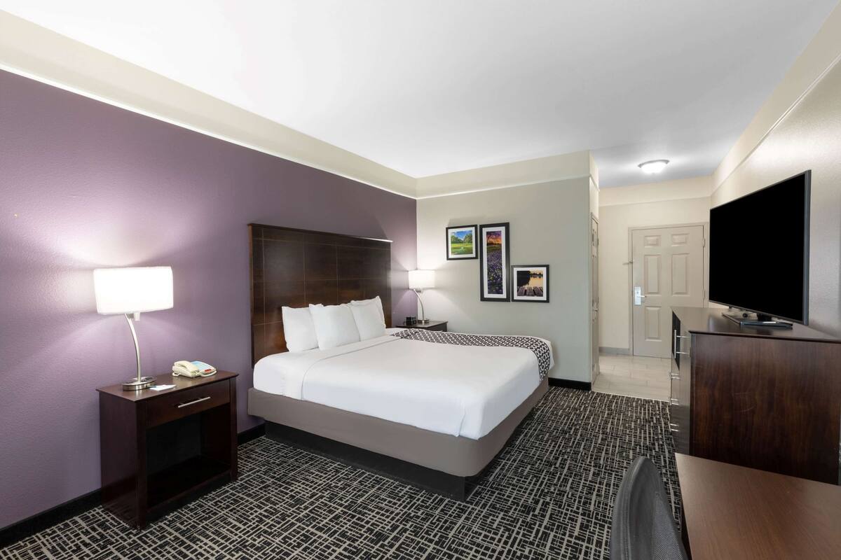 Una habitación de hotel con una cama grande, un escritorio con televisor de pantalla plana, una mesita de noche con lámpara y una puerta que da a otra habitación.
