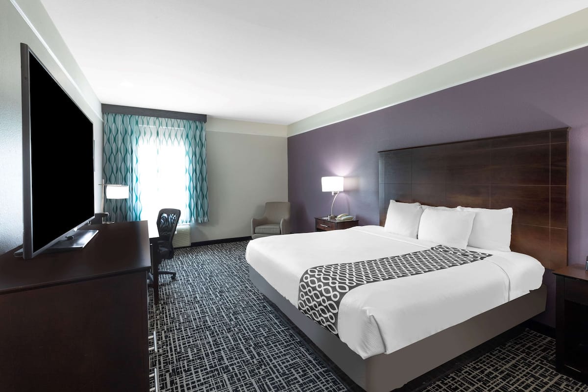 Habitación de hotel con una cama grande, un escritorio con televisión de pantalla plana, una silla, una lámpara y una alfombra estampada.