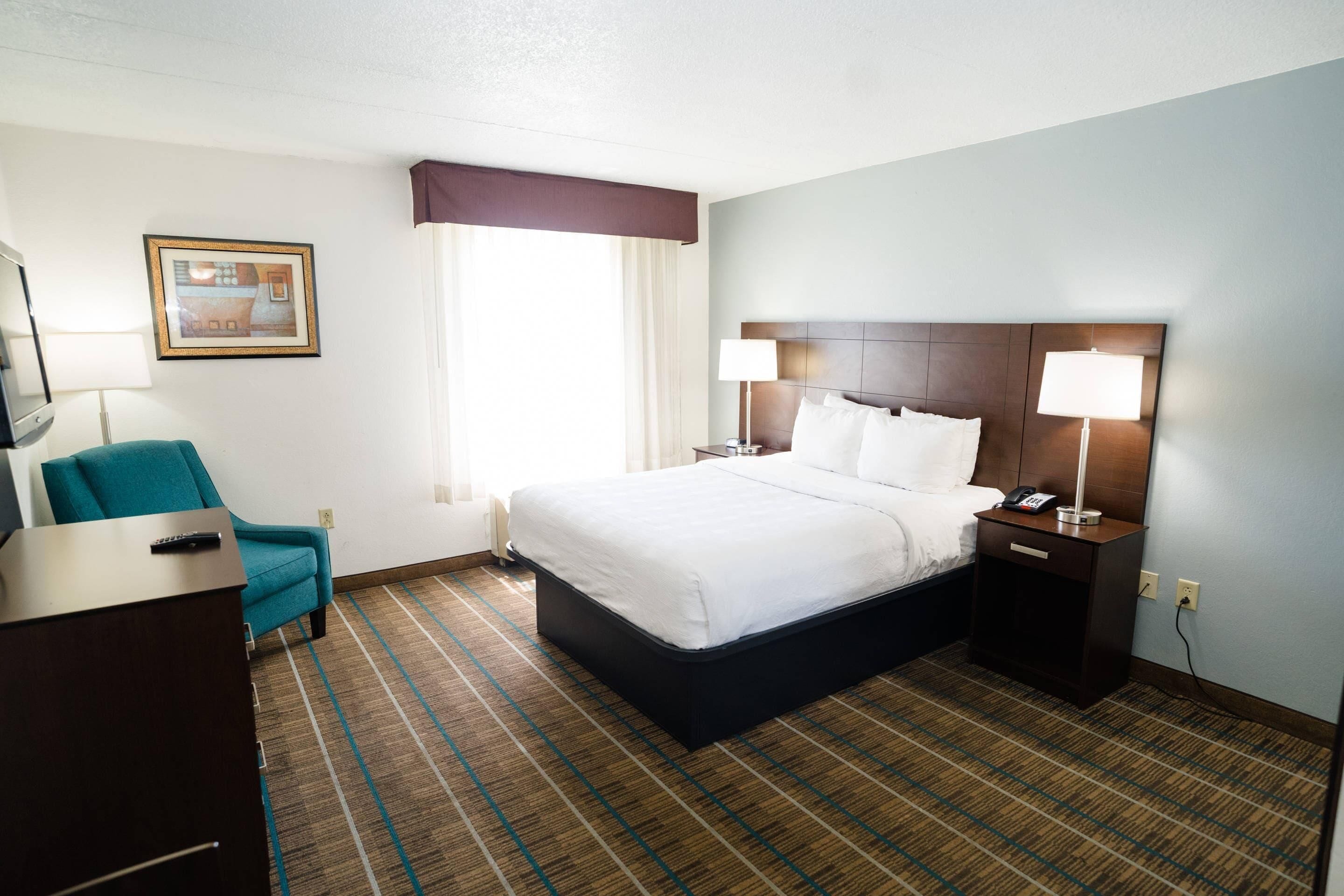 A hotel room with a bed, bedside lamps, a desk with a chair, a framed picture on the wall, and a window with curtains.