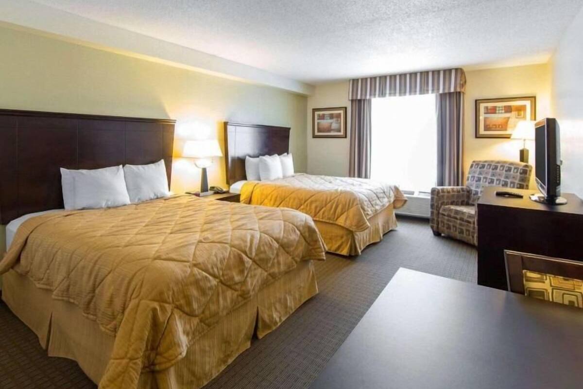 Habitación de hotel con dos camas, un escritorio con televisor, una silla y un ventanal con cortinas.