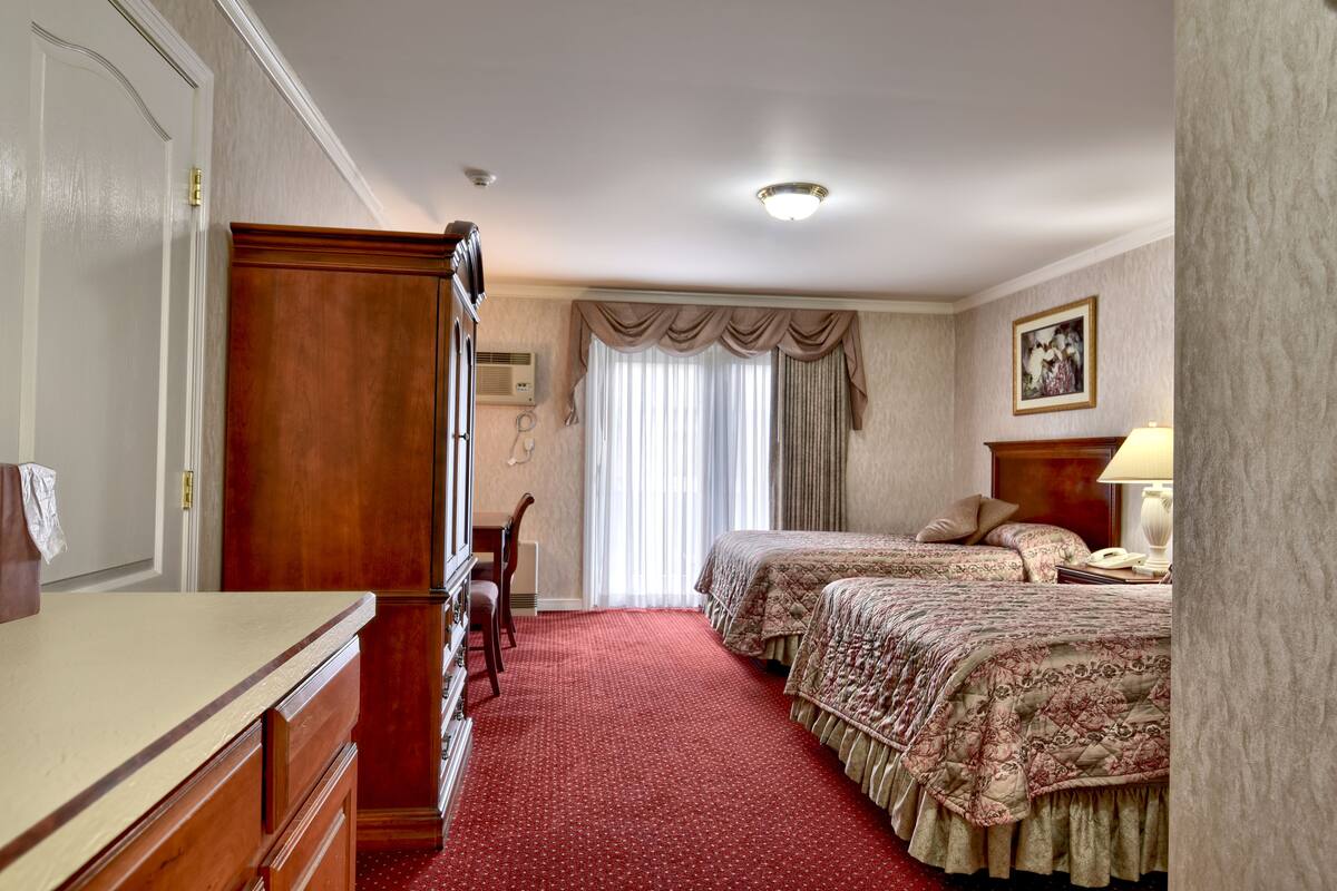 Habitación de hotel con cómoda de madera, dos camas con ropa de cama floral, alfombra roja, ventana grande con cortinas y un cuadro en la pared.