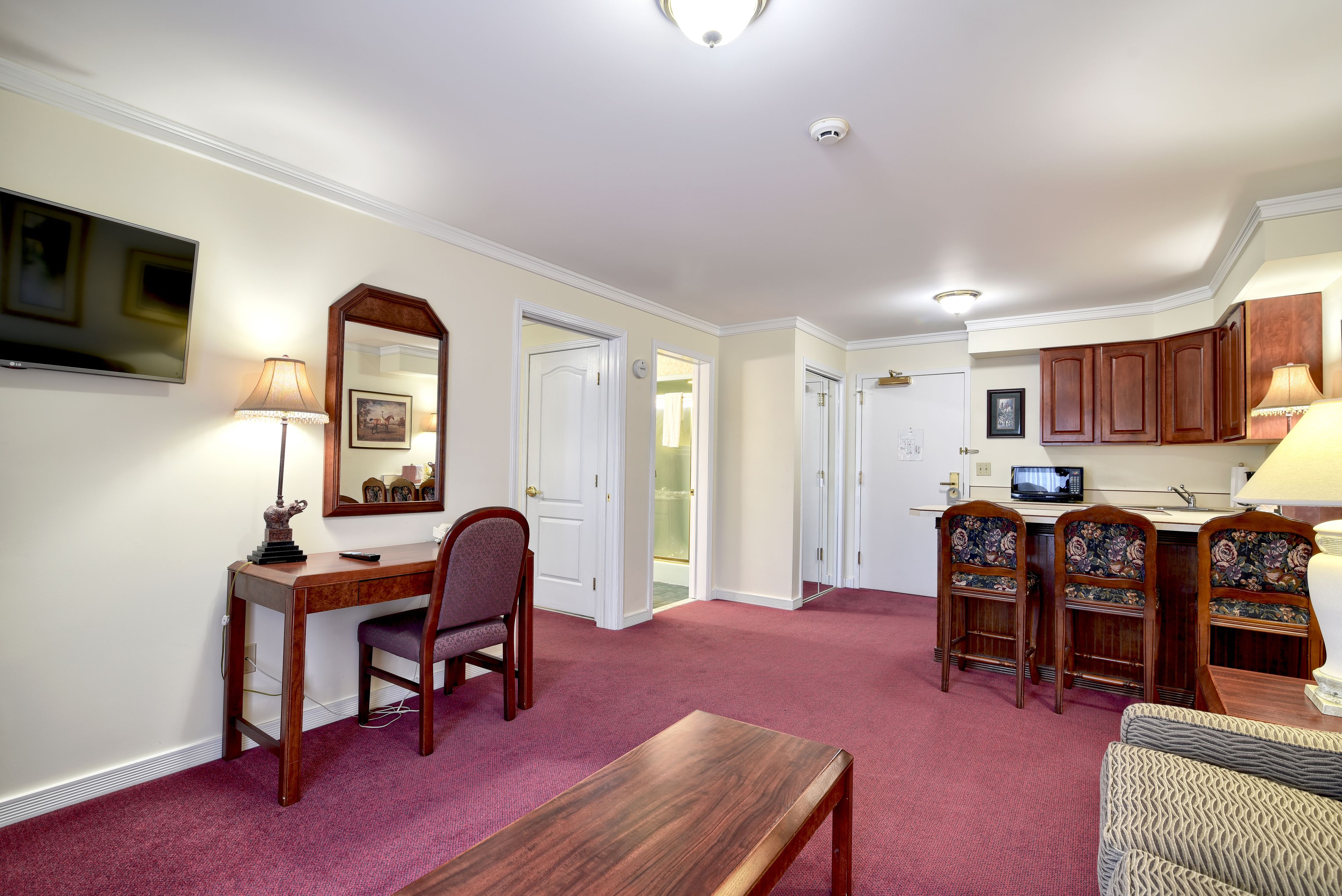 A hotel room with a desk, chair, and a small table. There is a television mounted on the wall, a lamp, and a mirror. The room has a carpeted floor and wooden cabinets.