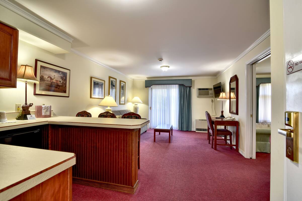 Habitación de hotel con barra de madera, un escritorio, una silla y una mesa de comedor. En la pared hay cuadros enmarcados, una ventana con cortinas y una puerta con un cartel.