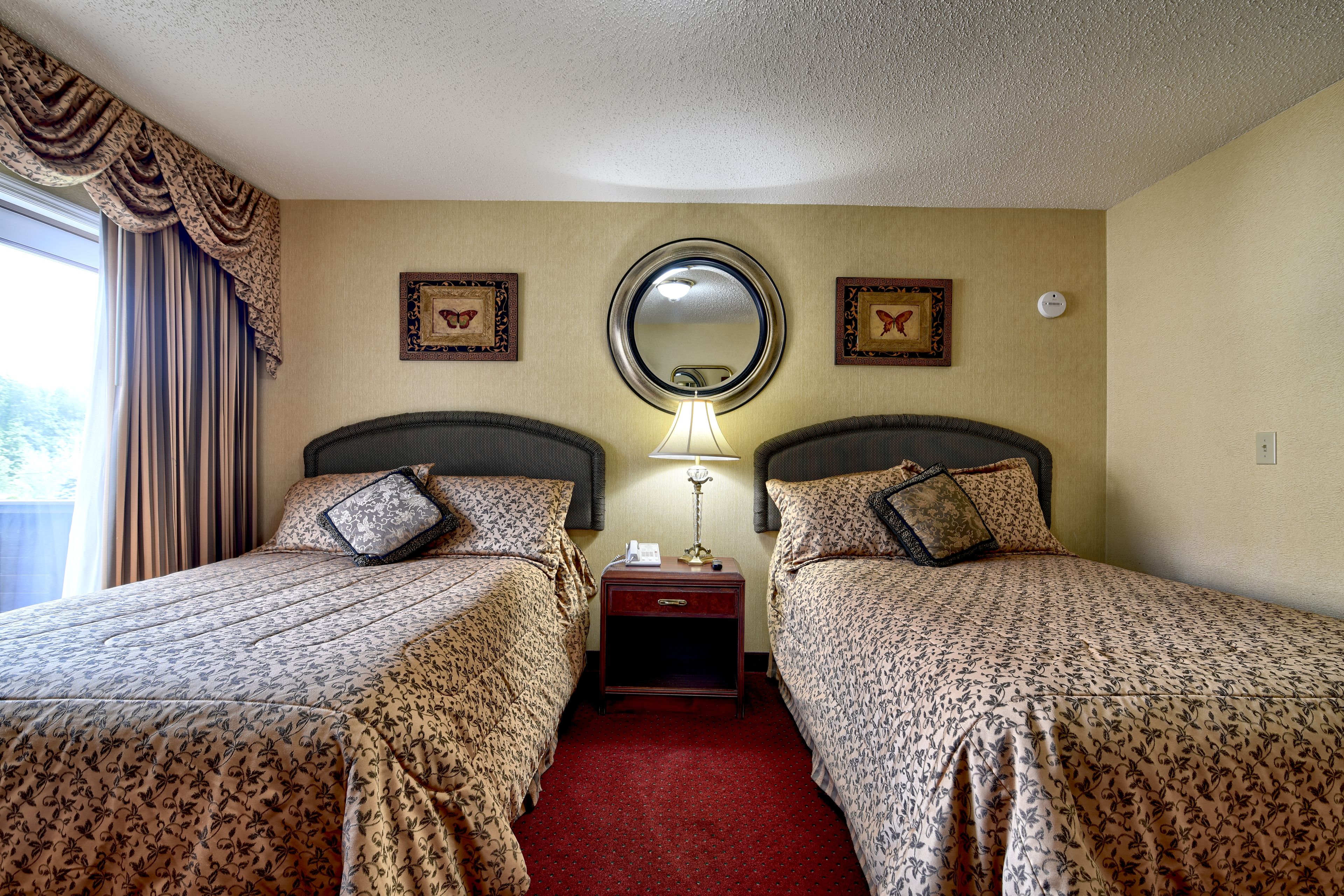 A hotel room with two beds, a nightstand, a lamp, a mirror, and framed pictures on the wall.