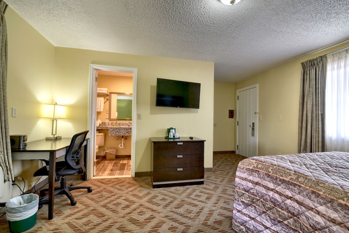 Habitación de hotel con cama, escritorio, silla, cómoda, televisor y baño visible a través de una puerta abierta.
