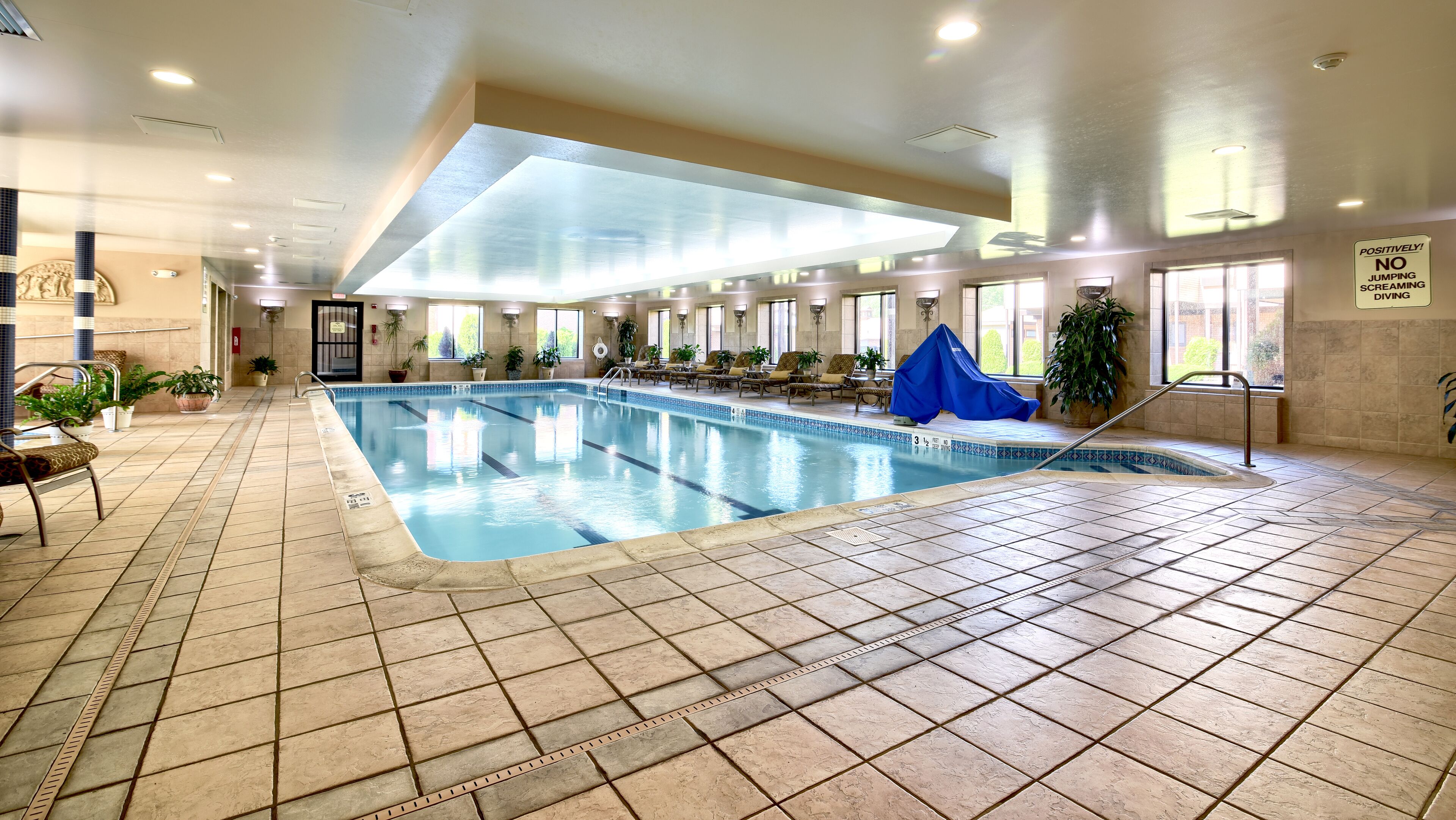 An indoor swimming pool with tiled flooring, lounge chairs, and potted plants.