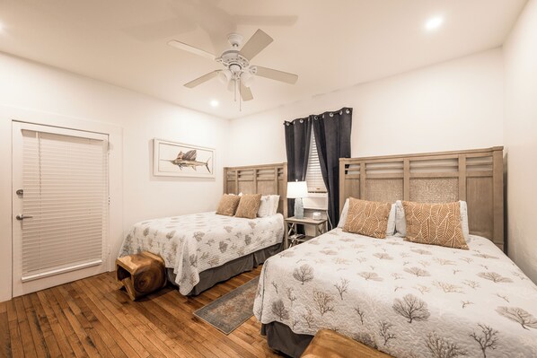 A bedroom with two beds, a ceiling fan, a desk with a lamp, and a window with curtains.