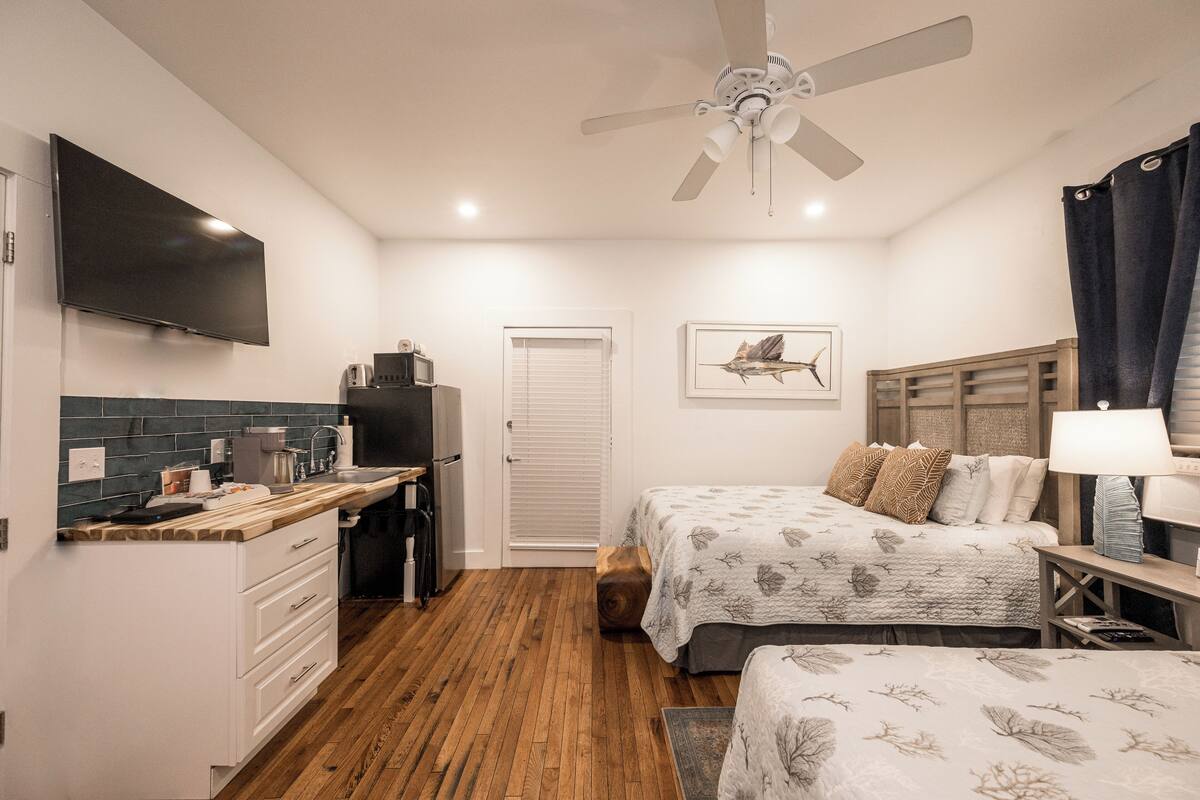 A bedroom with two beds, a ceiling fan, a TV, a desk with a coffee maker, and a window with blinds.