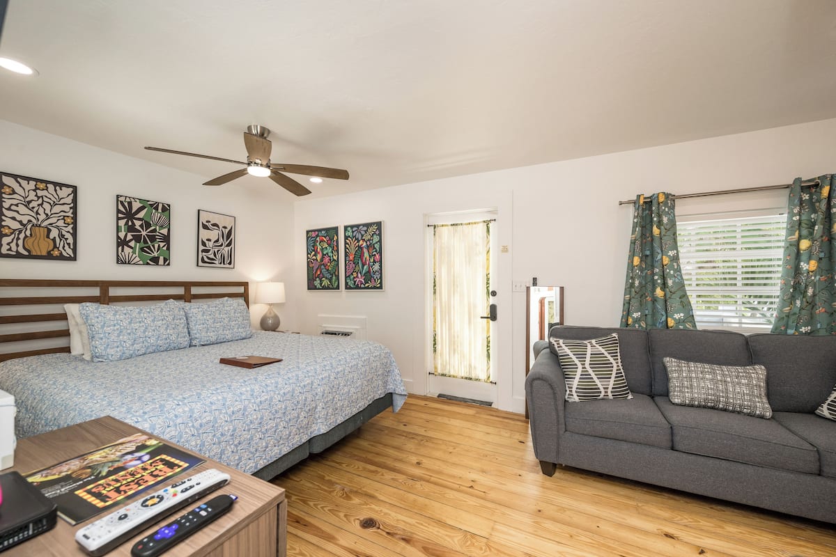 A bedroom with a bed, a gray sofa, wooden flooring, ceiling fan, artwork on the walls, and a window with curtains.