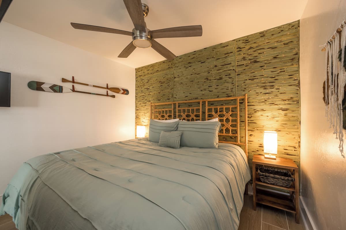 A bedroom with a bed, a ceiling fan, a wall-mounted TV, and a nightstand with a lamp.