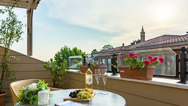 A round table with a book, a bottle, and a fruit platter on a balcony with potted plants and a view of a building with a tower. - Best Point Hotel (Istanbul)