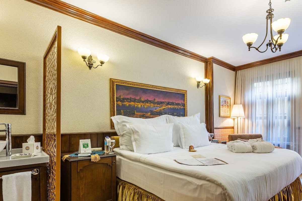 A hotel room with a large bed, a painting of a cityscape, a chandelier, and a desk with a lamp.