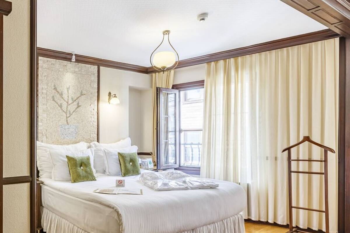 A hotel room with a large bed, a hanging light fixture, a window with curtains, and a coat rack.