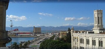 Un paisaje urbano costero con una torre destacada, un barco grande amarrado en un puerto y montañas al fondo.