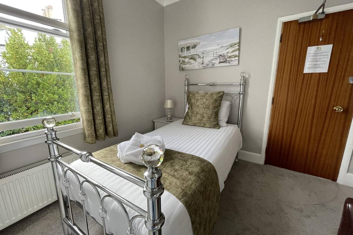 A bedroom with a bed, a window with curtains, a painting on the wall, and a door with a sign.