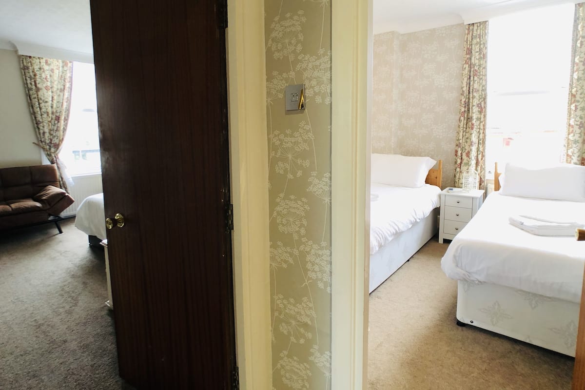 A hotel room with a bed, a bedside table, a nightstand, a lamp, a window with curtains, and a sofa in the adjacent living area.