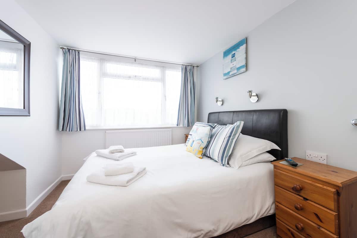 Una habitación de hotel con una cama, un aparador de madera, una ventana con cortinas y una lámpara fijada a la pared.