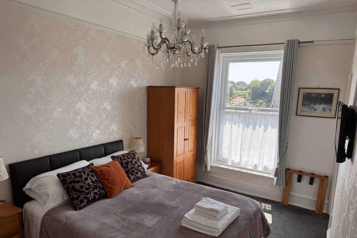 A bedroom with a bed, a wooden wardrobe, a chandelier, a window with curtains, and a painting on the wall.
