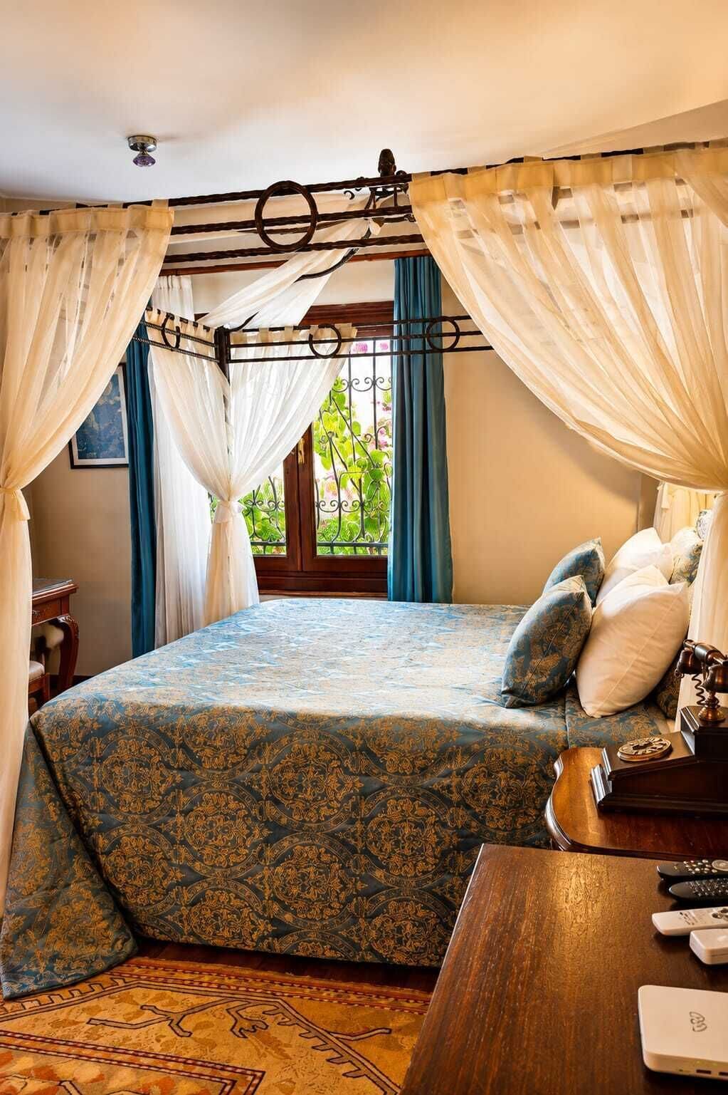 A bedroom with a canopy bed, a wooden nightstand, a lamp, and a view of greenery outside.