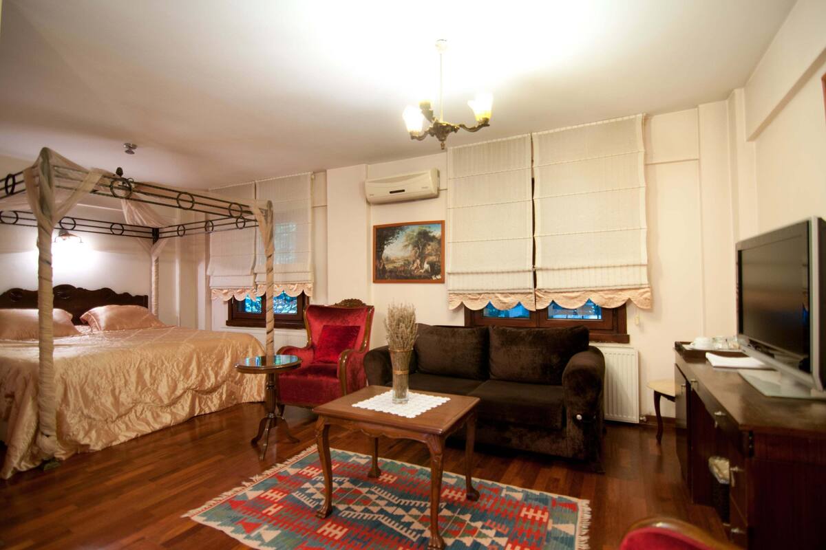 A bedroom with a four-poster bed, a red armchair, a sofa, a small table with a vase, and a television.