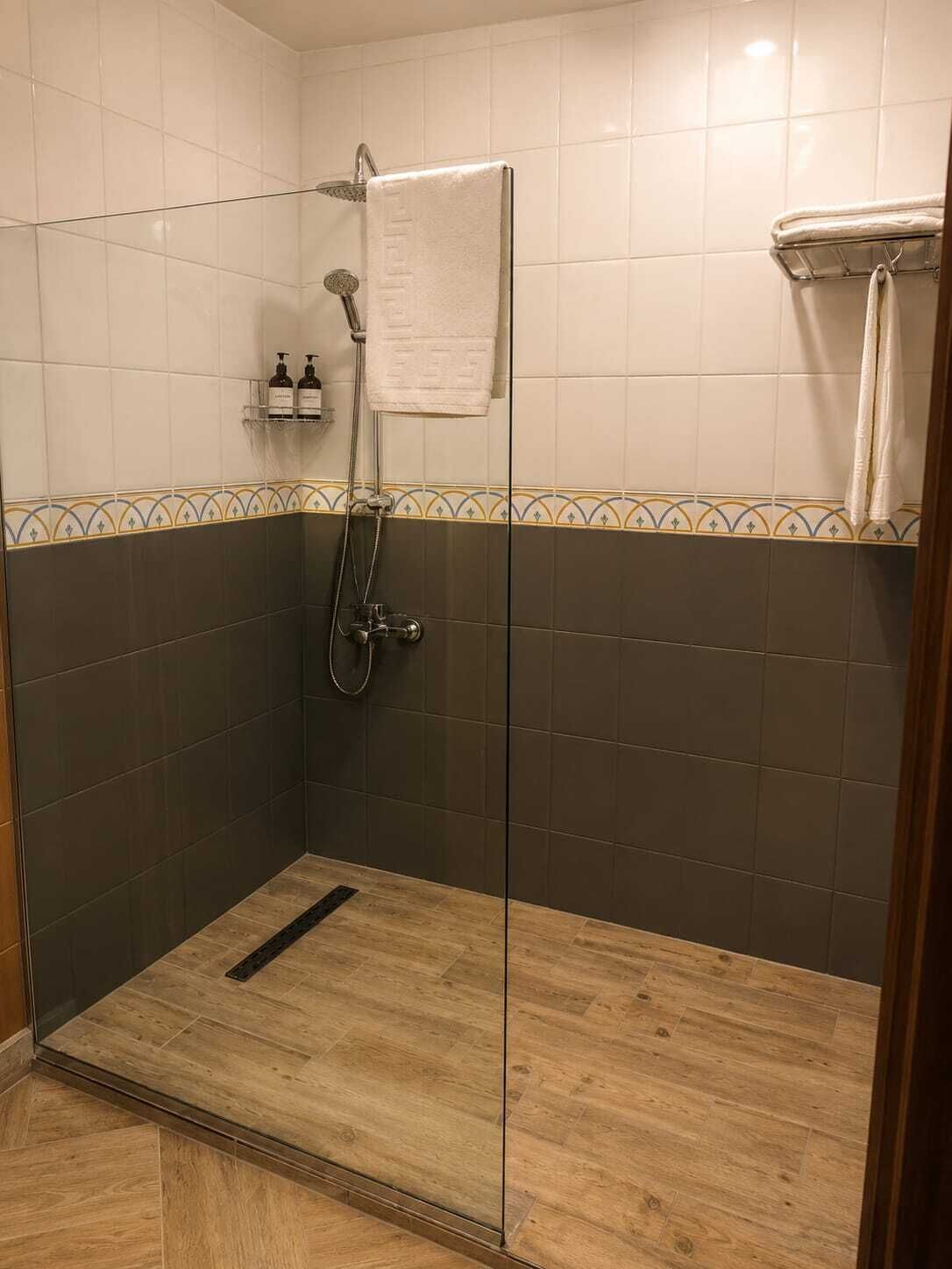 A glass-enclosed shower with a wooden floor, a towel hanging on the wall, and a shelf with toiletries.