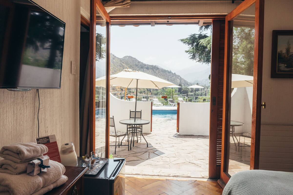 Una habitación con televisión, cama, mesa con silla, una pila de toallas y vistas a un patio exterior con sombrillas y piscina.