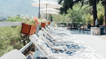 Relax in style with comfortable lounge chairs under shaded umbrellas, surrounded by lush trees and a breathtaking mountain view.