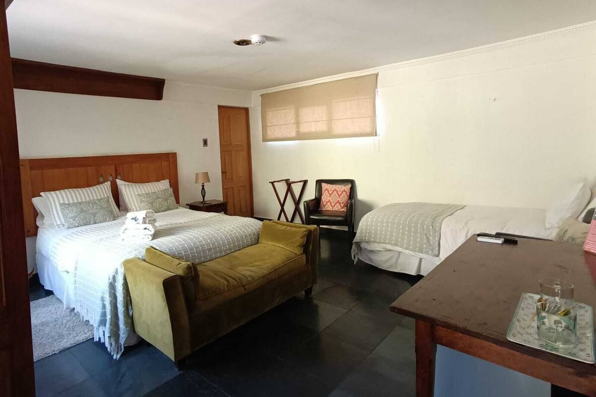 Un dormitorio con una cama grande, una cama más pequeña, una silla, una mesa y una ventana con persianas.