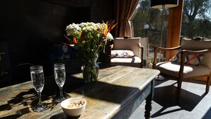 Una mesa de madera con dos vasos y un cuenco de nueces, un jarrón con flores, y vistas de un muro de ladrillo y una ventana con cortinas.