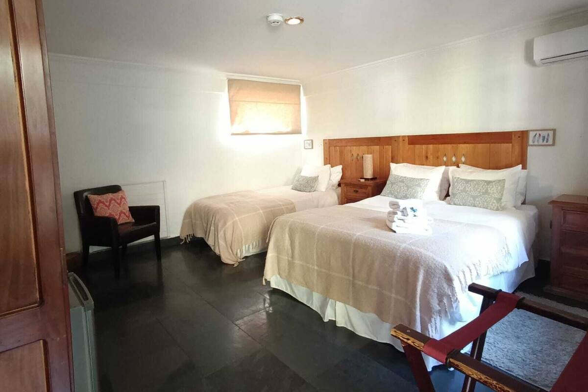 Habitación de hotel con dos camas, cabecero de madera, una ventana pequeña, una silla y un banco de madera.