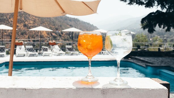 Dos vasos de bebidas sobre un borde de piscina, con sombrillas y sillones de descanso al fondo.