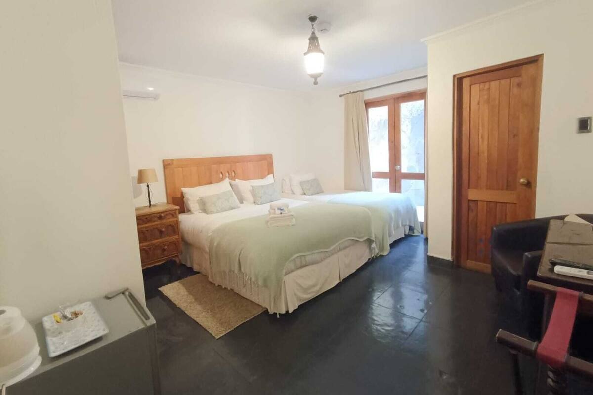 Un dormitorio con cabecera de madera, una cama con ropa de cama blanca, una mesita de noche con lámpara, una puerta de madera y una pequeña mesa con una caja.