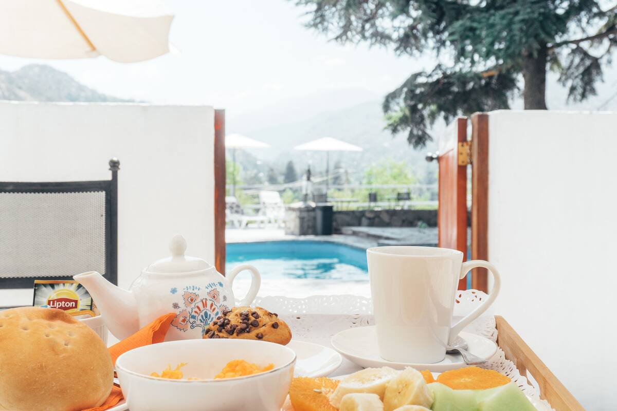 Bandeja de desayuno con tetera, taza, plato y bollería, con vistas a una piscina y vegetación.