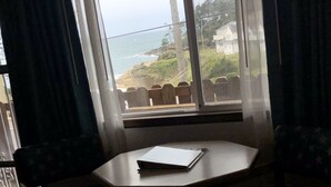 A room with a view of the ocean through a window with curtains. There is a small table with a notebook on it.