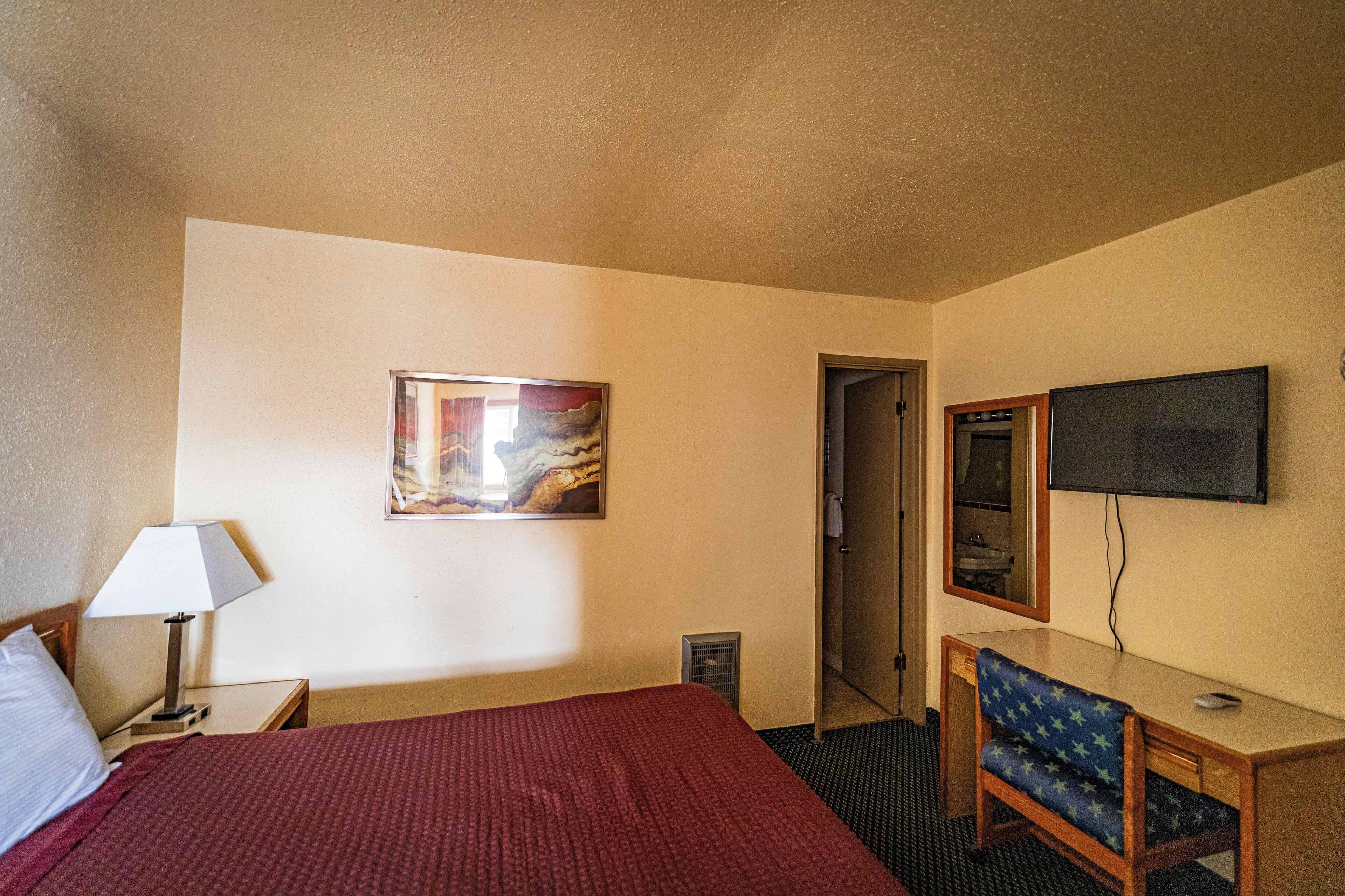 Habitación de hotel con cama, escritorio, silla, televisor, un cuadro y una puerta.
