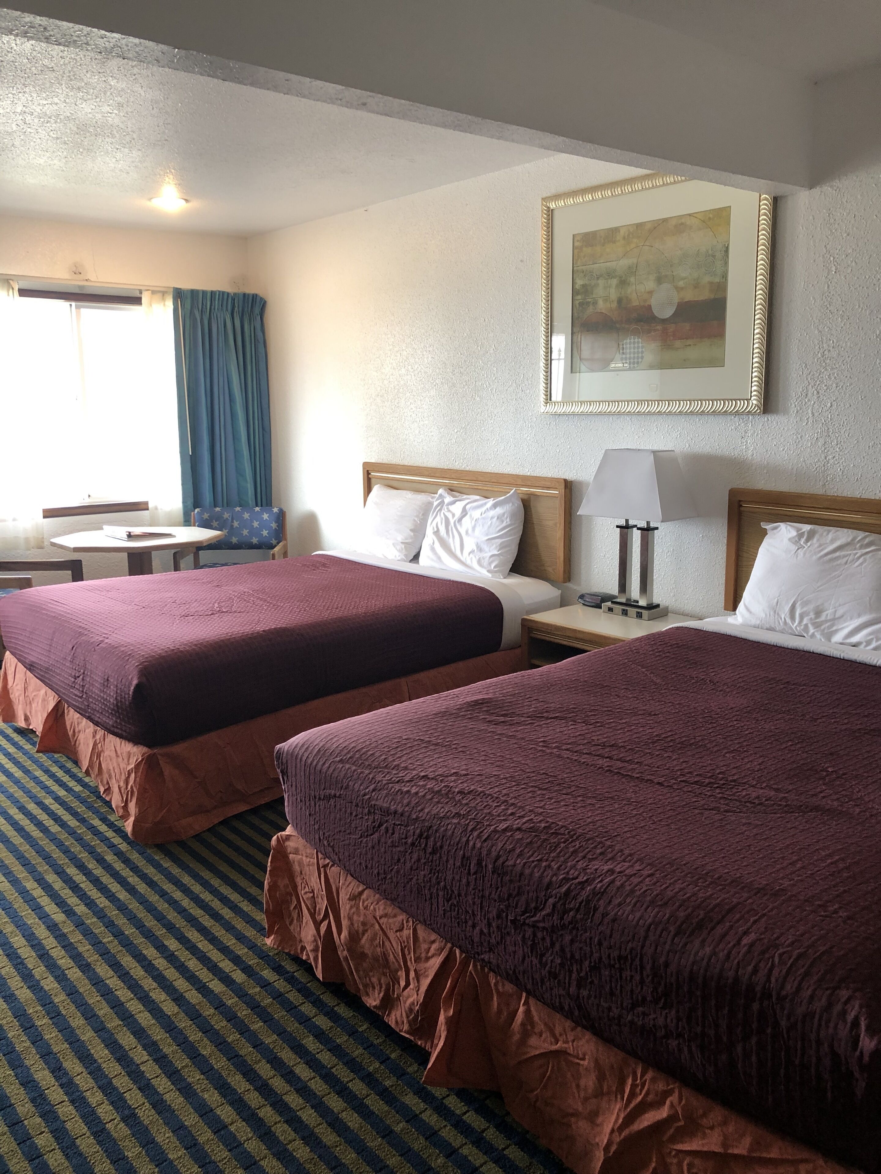 A hotel room with two beds, a small table, a chair, a lamp, and a painting on the wall.