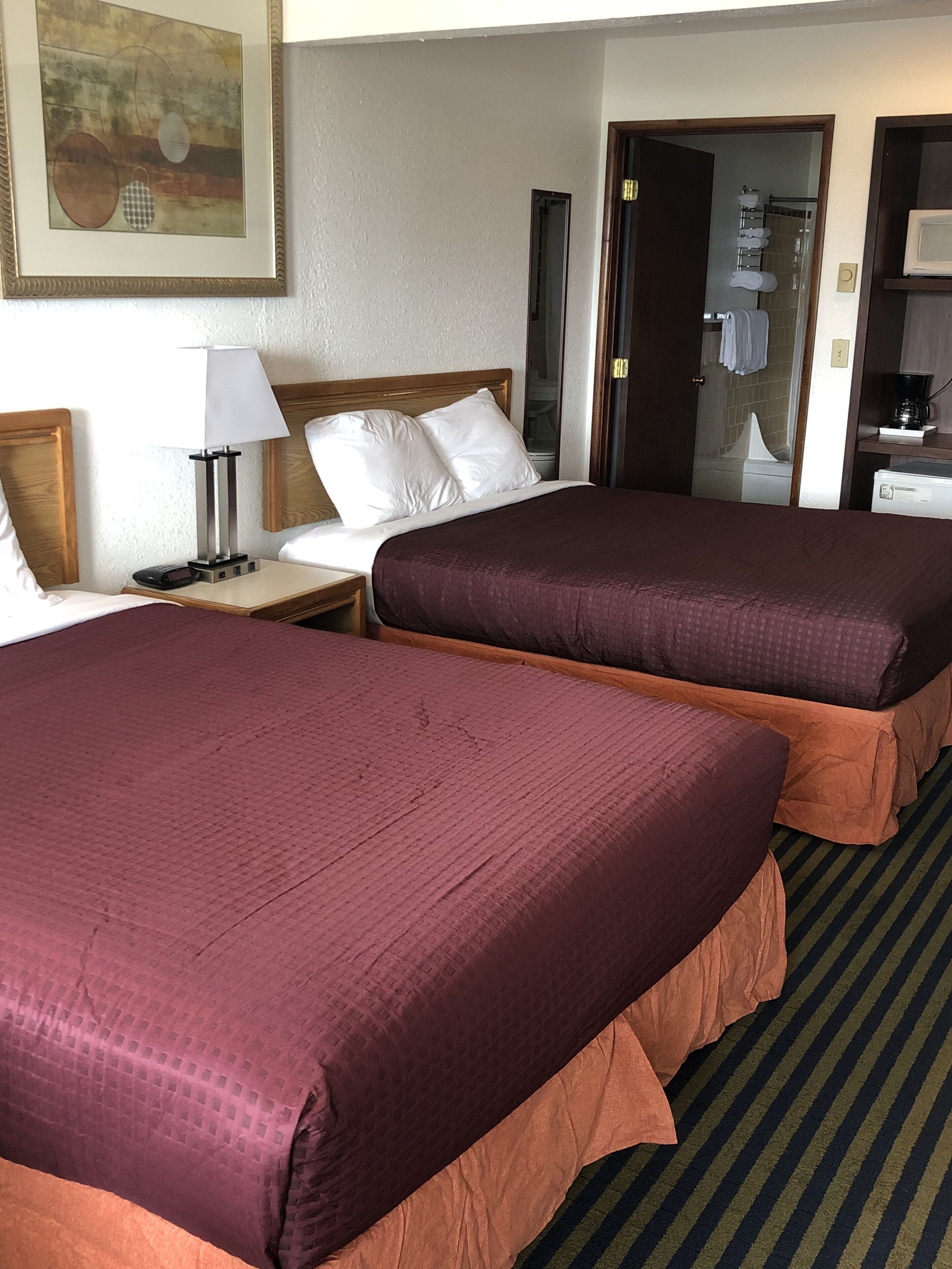 A hotel room with two beds, a nightstand with a lamp, a framed picture on the wall, and a view of the bathroom through an open door.