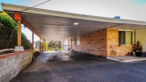 A covered parking area with a brick building, a wooden chair, and a window with a curtain.