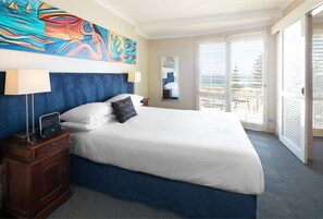 A hotel room with a large bed, a nightstand with a lamp, a painting on the wall, and a view of the ocean through the sliding doors.