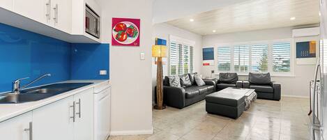 A modern kitchen with white cabinets, a blue backsplash, and a stainless steel sink. There is a black leather sofa set and a coffee table in the living area.