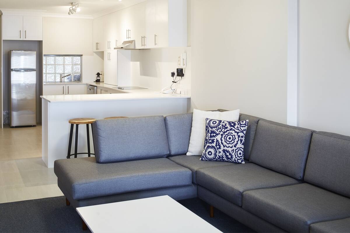A modern living room with a grey sectional sofa, a white coffee table, and a kitchen area in the background.