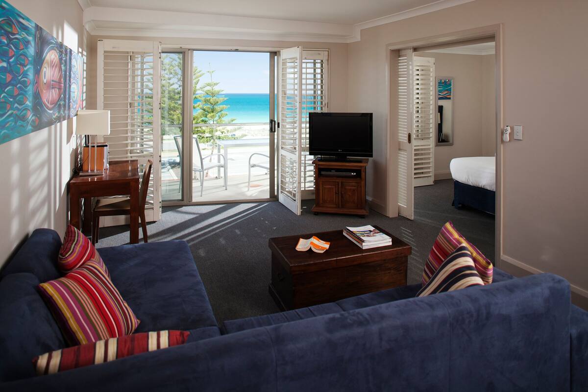 A living room with a blue sofa, a coffee table with books, a TV, and a balcony with a view of the ocean.