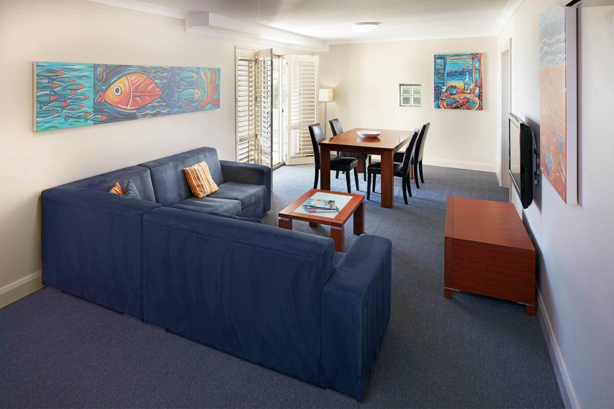 A living room with a blue sofa, a wooden coffee table, and a dining area with a table and chairs. There are paintings on the walls and a TV on the wall.