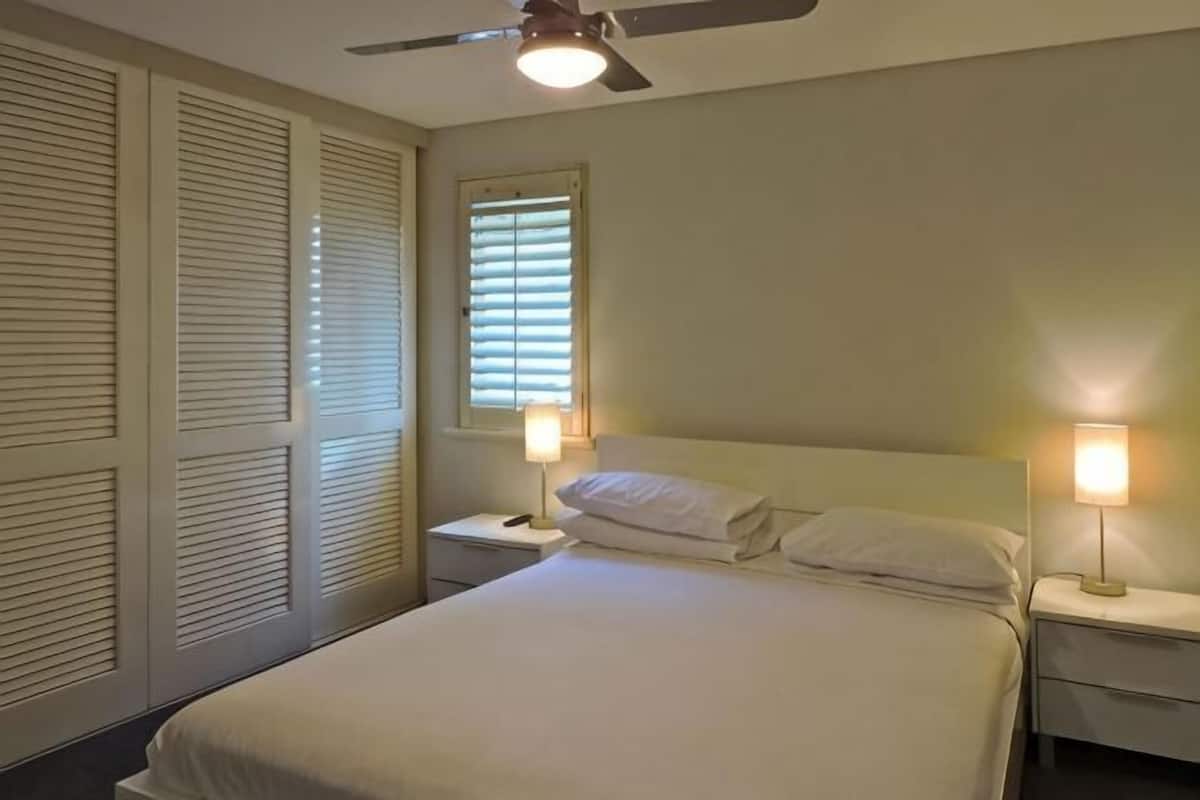 A bedroom with a bed, two bedside tables, a ceiling fan, a window with blinds, and a nightstand with a lamp.