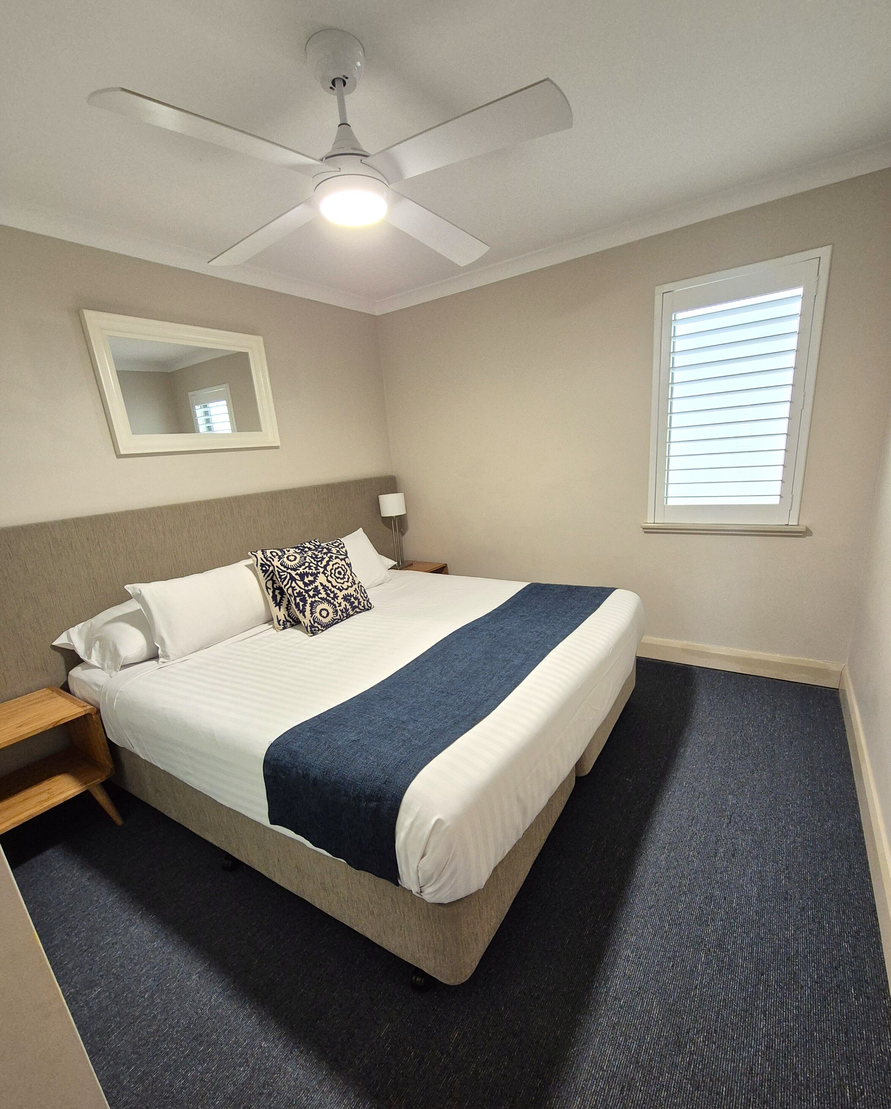 A hotel room with a bed, a ceiling fan, a nightstand, a mirror, a window with blinds, and a carpeted floor.